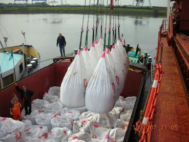 Rozładunek worków big bag z cementem 42,5 I za pomocą dźwigu z barki na nabrzeże, w tle widoczny port i rzeka.